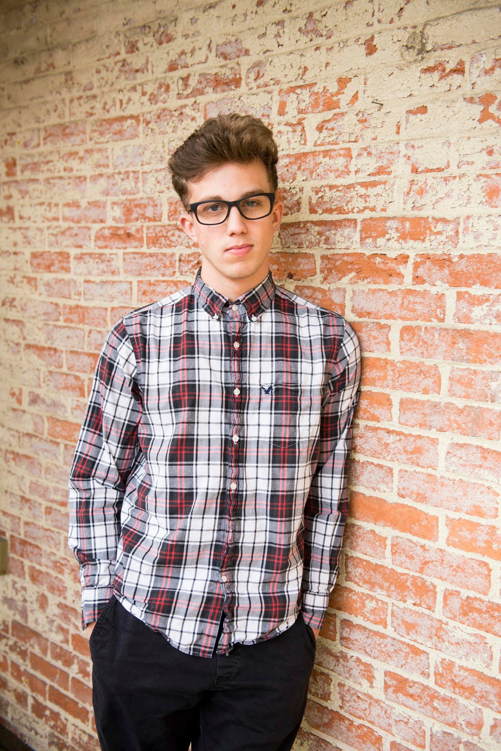 Trendy young man in glasses, plaid shirt, posing confidently against a brick wall.